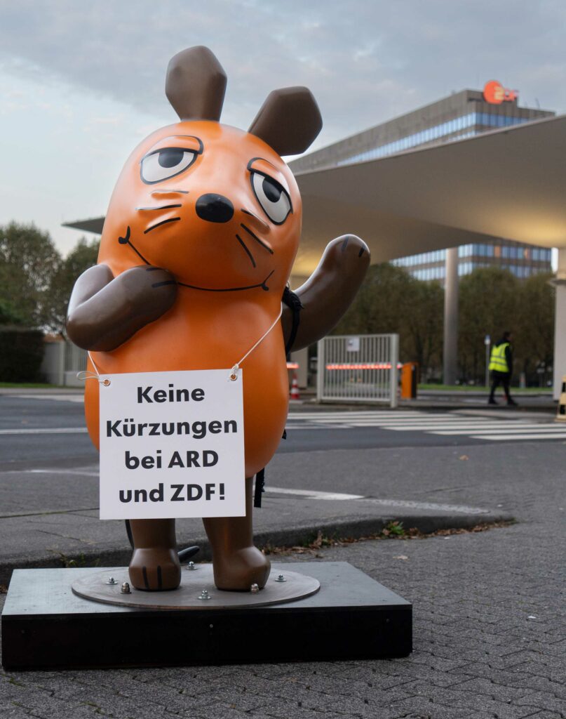 Die Maus steht vor dem ZDF in Mainz. Sie trägt ein Schild mit der Aufschrift "Keine Kürzungen bei ARD und ZDF!"