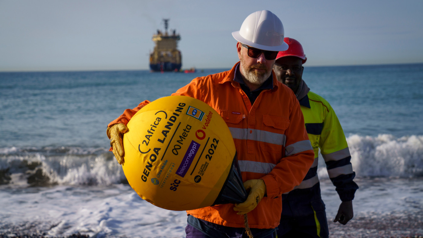 Zwei Ingenieure im Auftrag von Meta bzw. des Projektes 2Africa tragen eine goldene Boje an den Strand. Sie markiert den Anschluss des Glasfaserkabels an das Festland.