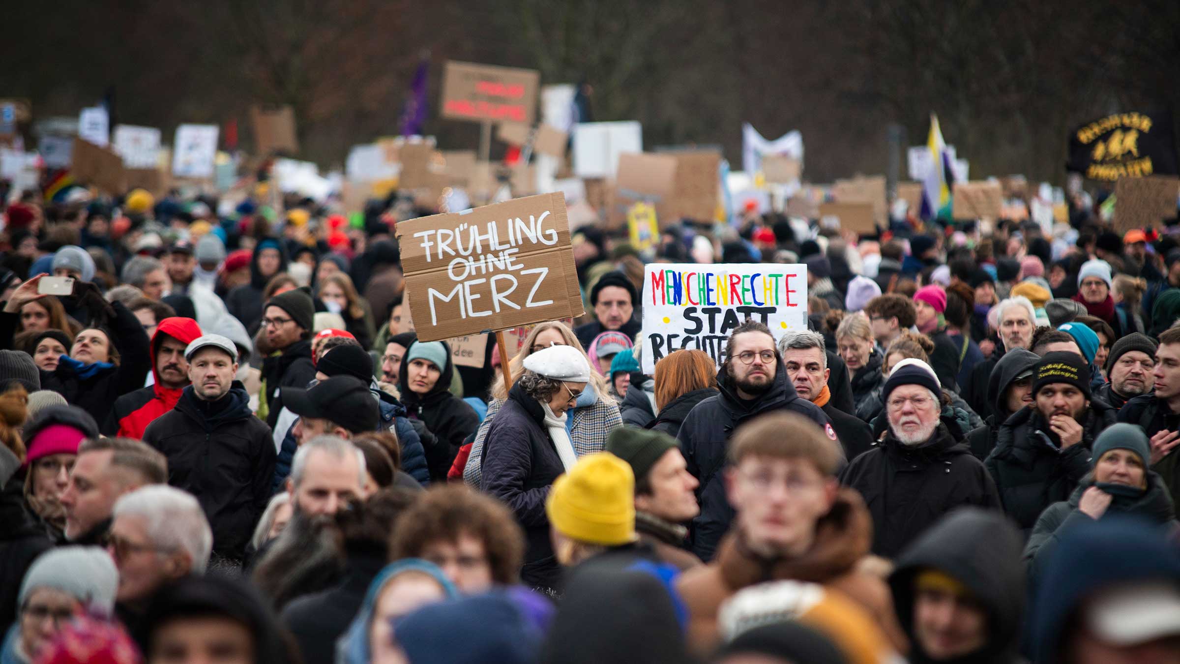 Eine Gruppe von Menschen demonstriert in Berlin gegen die Zusammenarbeit von Merz mit der AfD. In der Mitte des Bildes hält eine Person ein Schild hoch, darauf steht: Frühling ohne Merz.