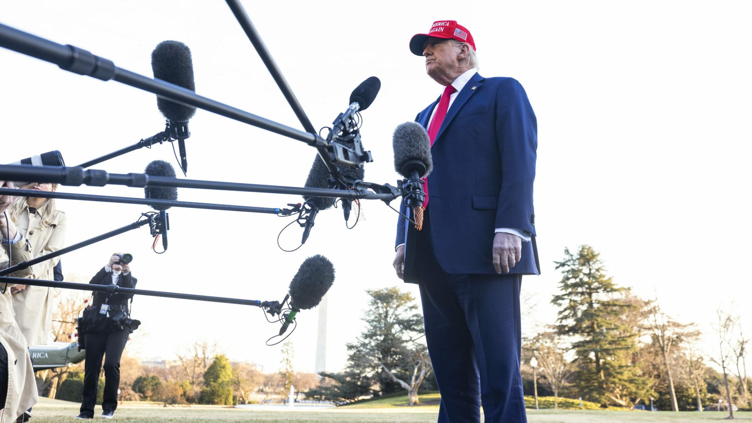 US-President Donald Trump in einem blauen Anzug, roter Krawatte und roter Baseballkappe steht im Freien vor mehreren Mikrofonen, die auf ihn gerichtet sind. Im Hintergrund sind Bäume, das Washington Monument und Fotograf*innen zu sehen.