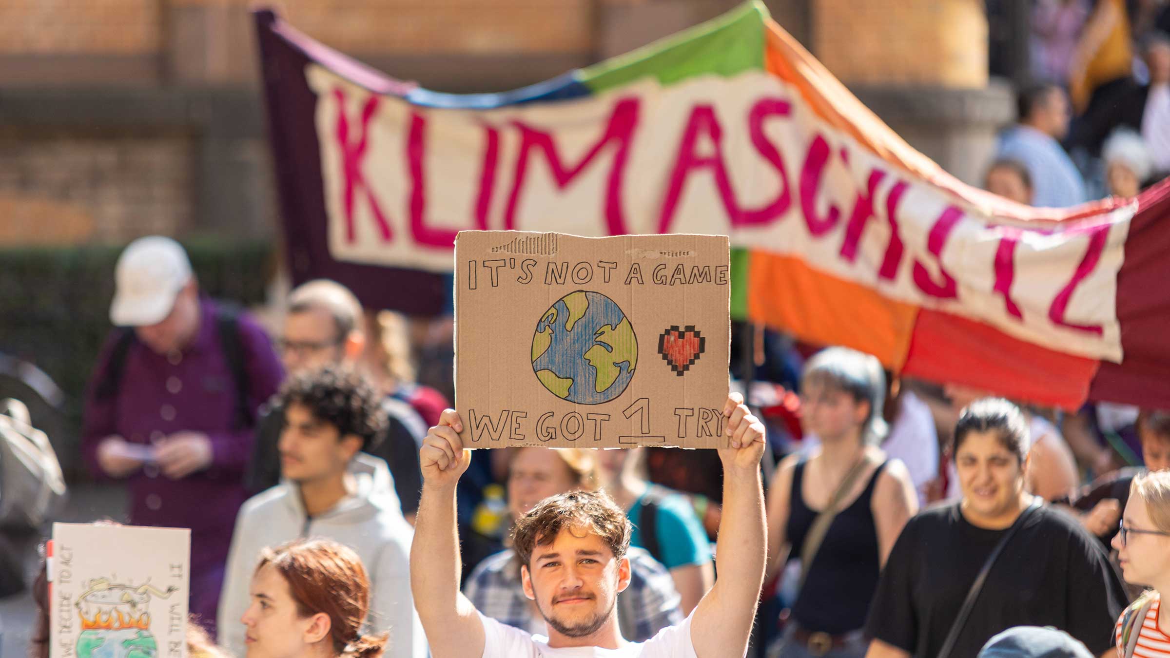 Demo von Klimastreik von Fridays For Future in Bonn am 20.09.2024. Im Hintergrund ein Banner mit dem Wort „Klimaschutz“. Im Vordergrund hält ein Mann ein Schild in die Höhe. Darauf steht. „It’s not a game. We got 1 try.“ Übersetzt heißt dies: Es ist kein Spiel, wir haben einen Versuch.