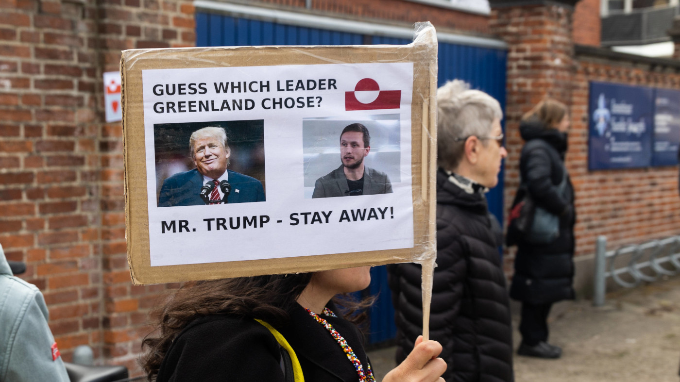 Eine Person hält bei einem Protest ein selbstgebasteltes Schild vor ihr Gesicht. Auf dem Schild steht oben, auf Englisch: "Rate mal, welches Oberhaupt Grönland gewählt hat?" Daneben ist die Flagge von grönland abgebildet. Darunter sind zwei Fotos: Eins von Donald Trump, dem Präsidenten der USA. Daneben ein Foto von Jens-Frederik Nielsen, dem kürzlich gewählten Regierungschef von Grönland. Unter den Fotos steht der Schriftzug: "Mr Trump - Bleiben Sie weg!"