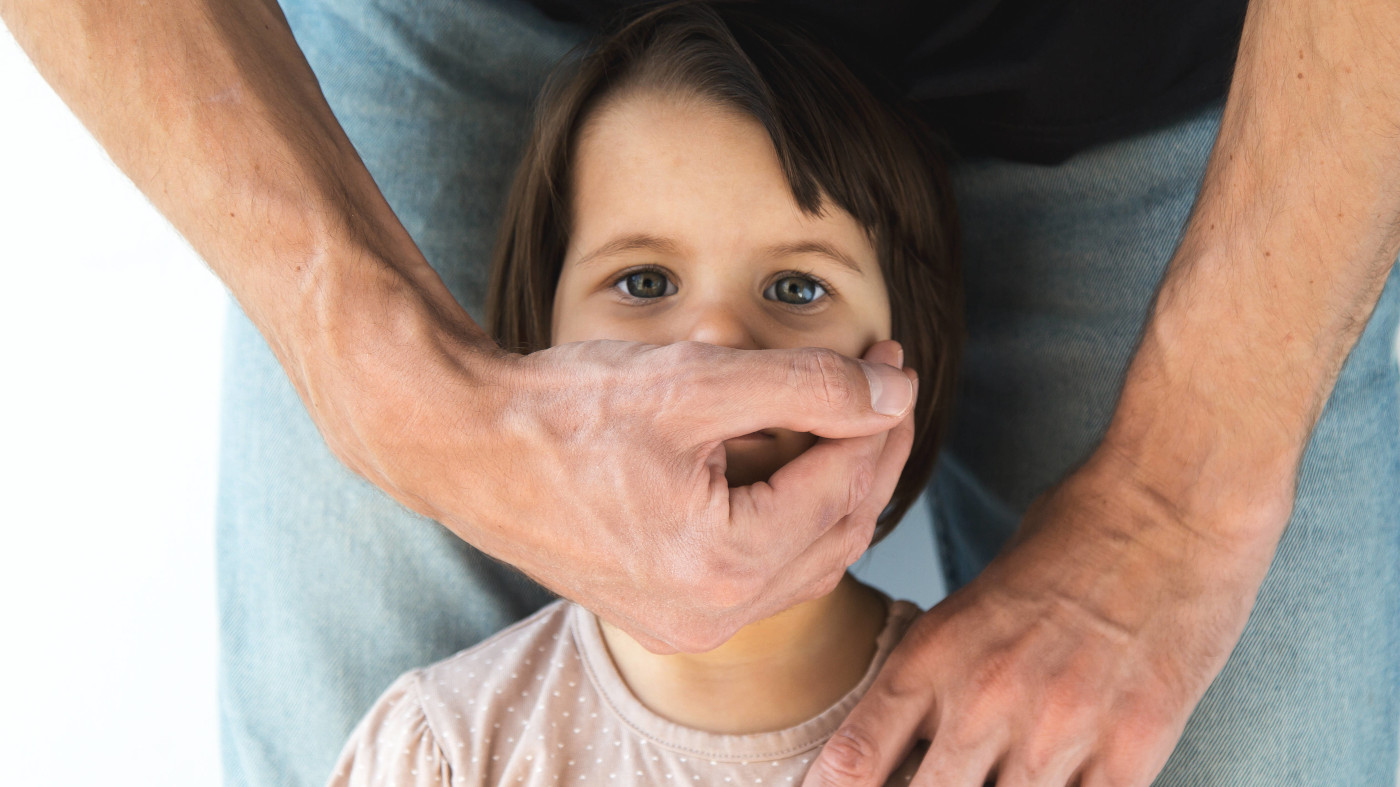 Nahaufnahme: Eine männliche Hand bedeckt den Mund eines etwa 3-jährigen Mädchens. Der Mann steht hinter dem Mädchen, es sind aber nur seine Hose, ein Teil des T-Shirts und seine Arme zu sehen.